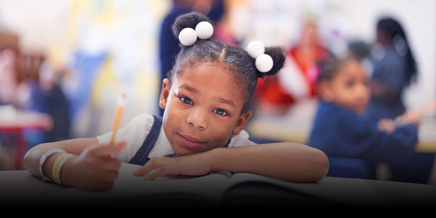 Young student writing in a notebook at a classroom desk.