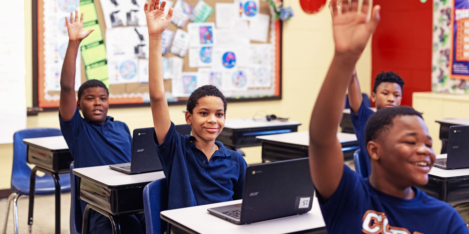 Students at desks with laptops raising their hands in class.